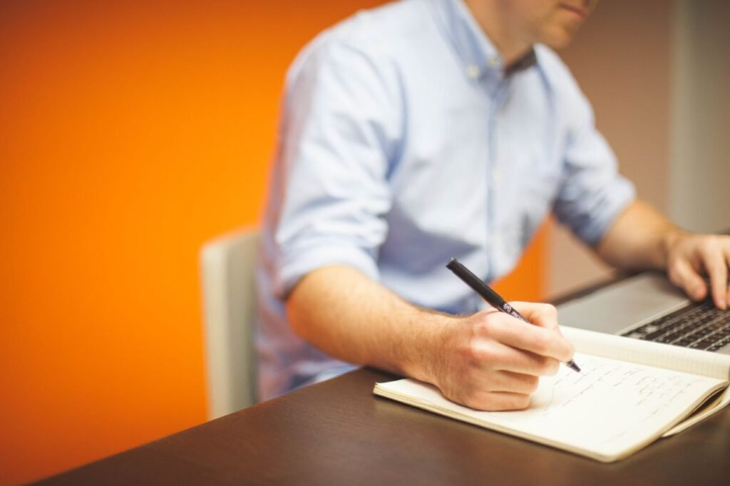 A man sits in modern office, writing notes with a laptop open.