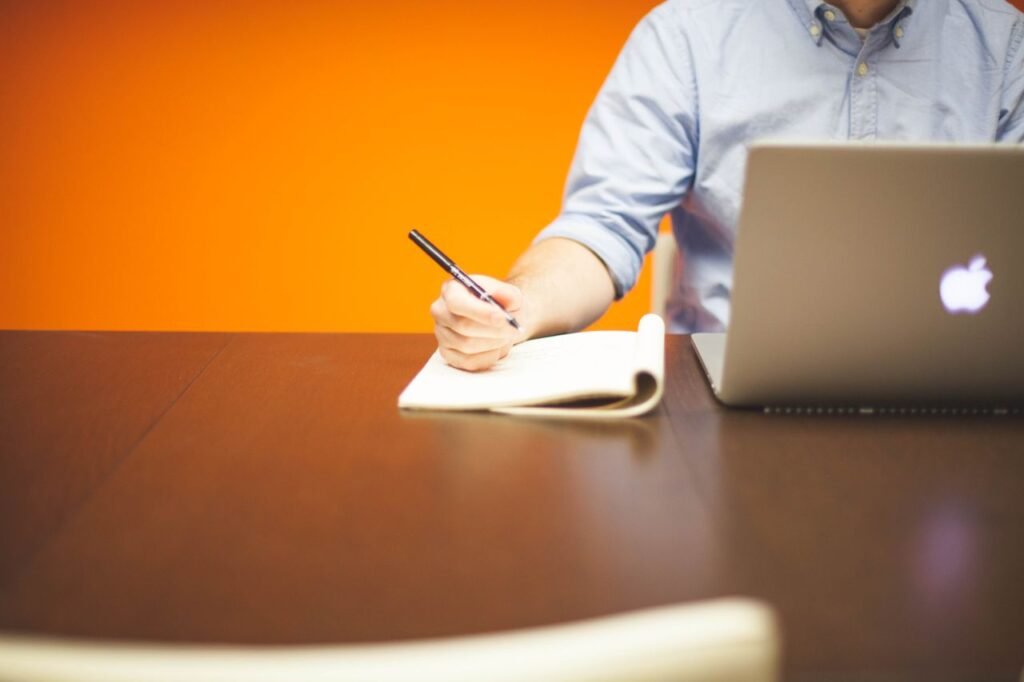 Adult writing notes in a notebook beside a laptop on a wooden desk with orange background.