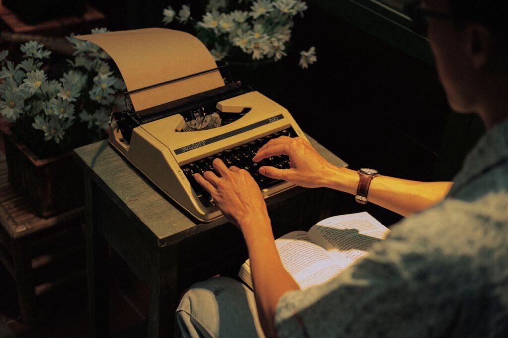 A person typing on a vintage typewriter indoors, surrounded by flowers, with a warm and nostalgic atmosphere.