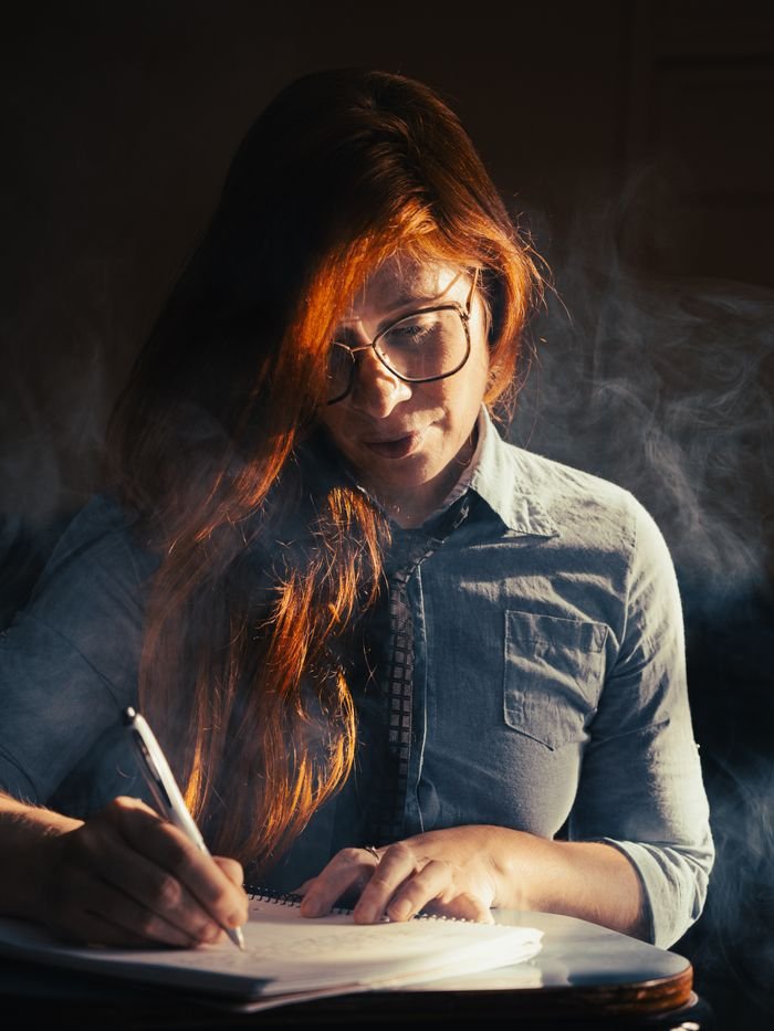 A woman with red hair writes in a notebook, surrounded by smoke, under warm lighting.