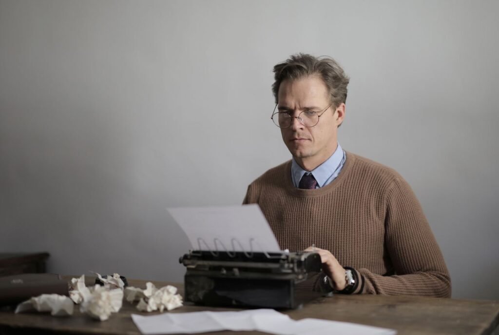 Man concentrating on writing at a desk with a vintage typewriter surrounded by crumpled paper.