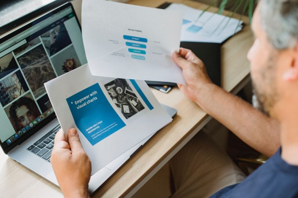 Crop anonymous male with beard looking at documents with graphic charts while sitting at table with open laptop