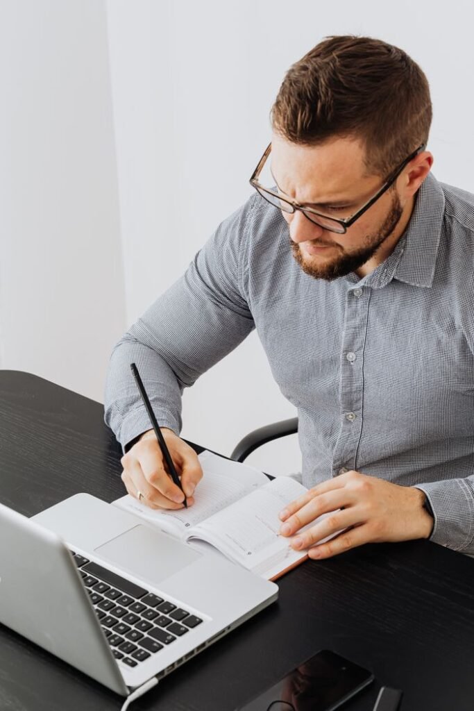 A man writing in a notebook while using a laptop, featuring an office setting for productivity and business.