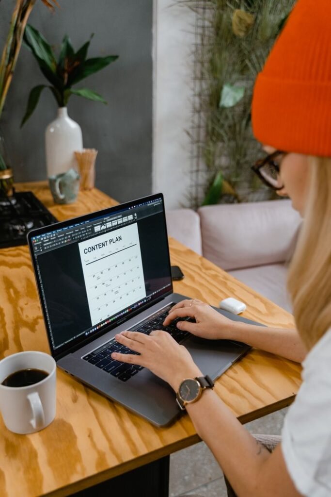 A woman wearing a knit hat types on a laptop at a wooden table with a cup of coffee indoors.