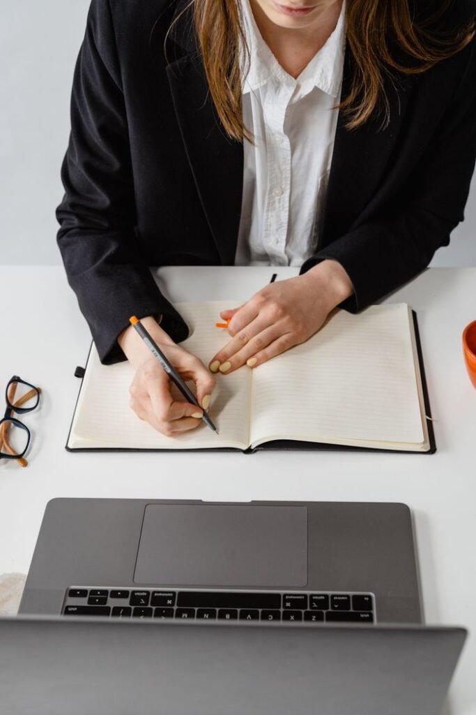Overhead view of a professional woman in a black blazer writing in a notebook at her desk.